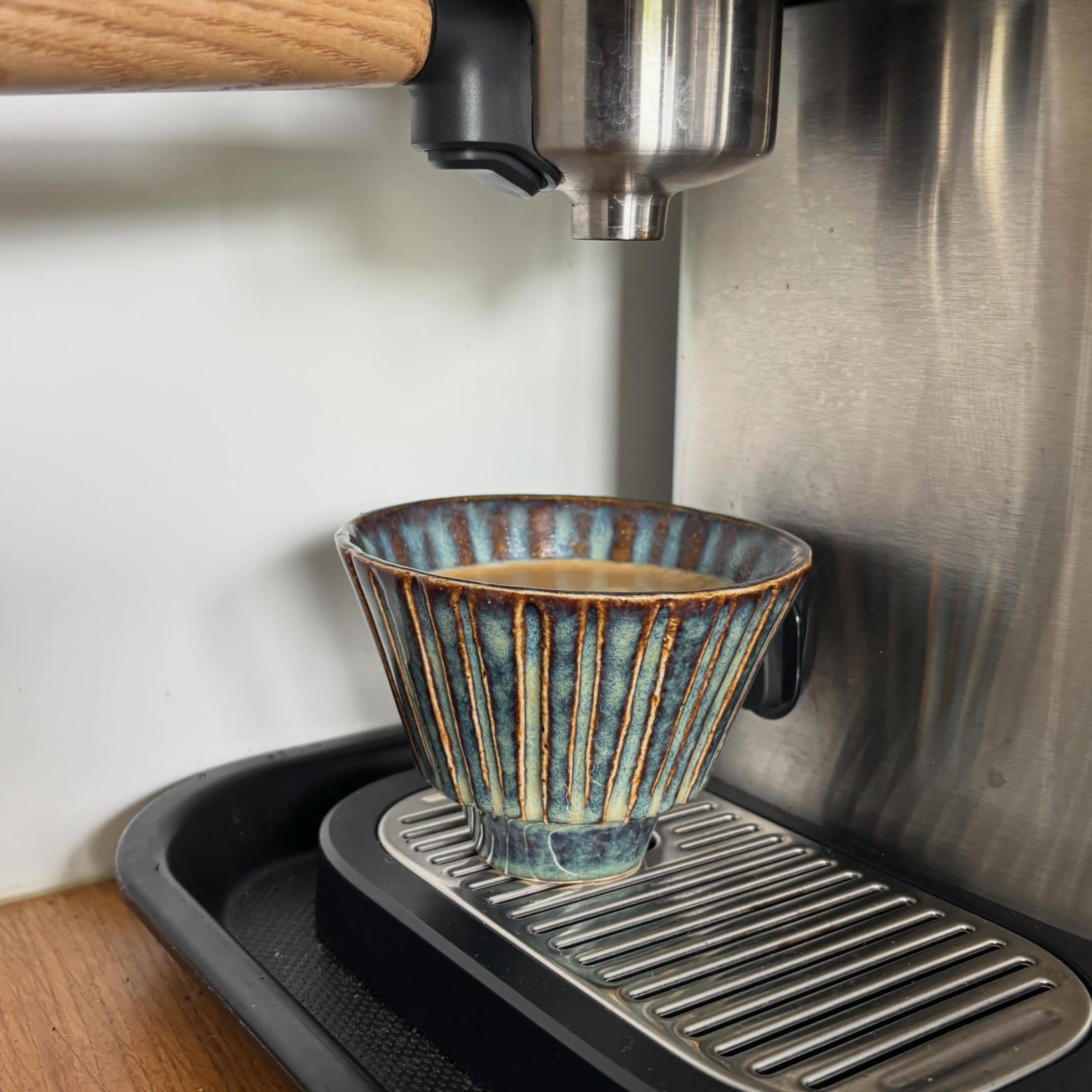 Geribbeld kopje in blauwgroen en bruin glazuur onder een espressomachine, gevuld met koffie