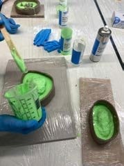 Groen mallenmateriaal wordt in bakjes van klei gegoten, met handschoenen en spuitbussen ernaast