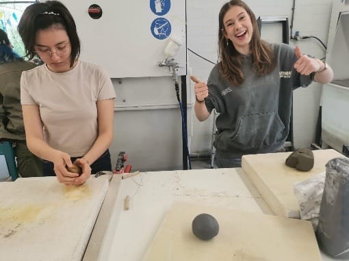 Twee studenten aan een werktafel met klei, een van hen steekt lachend haar duimen op