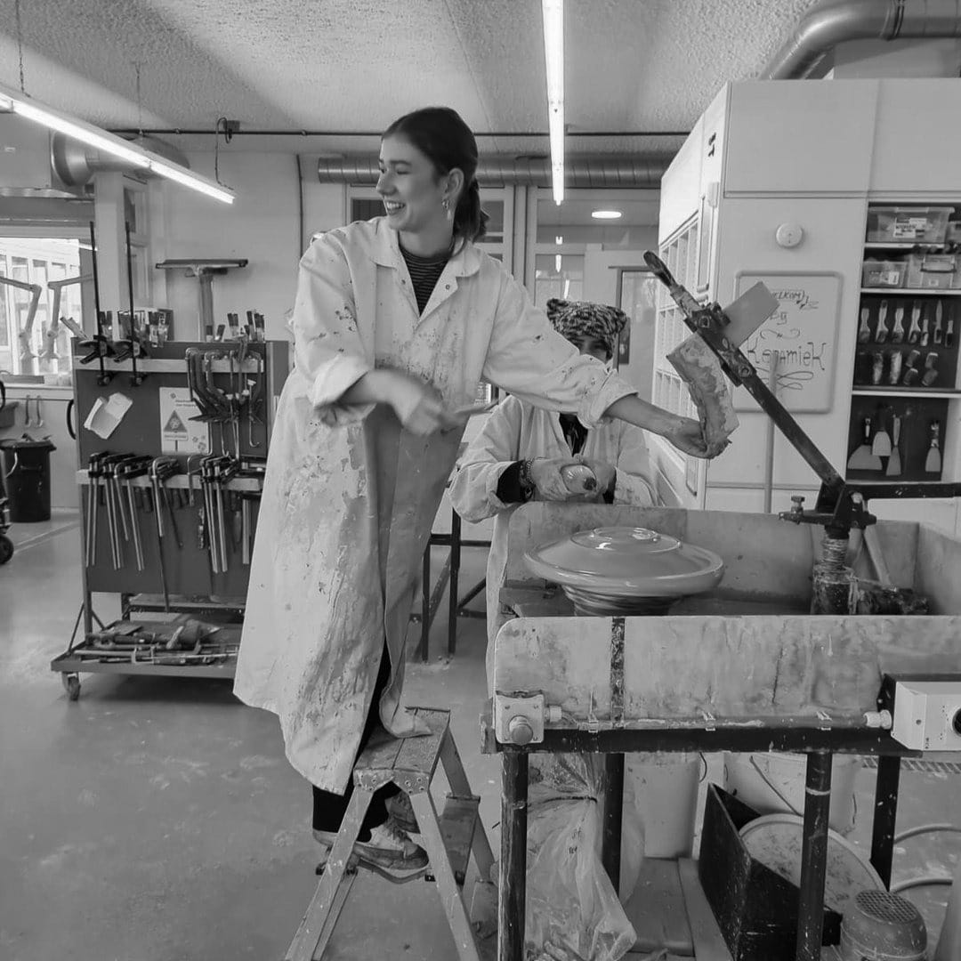 Lachend in een witte werkjas bij de draaischijf in de werkplaats, in zwart-wit