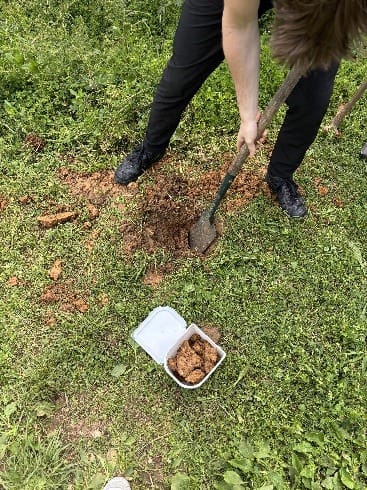 Met een schep wordt klei uit de grond gestoken, naast een bakje met verzamelde klei